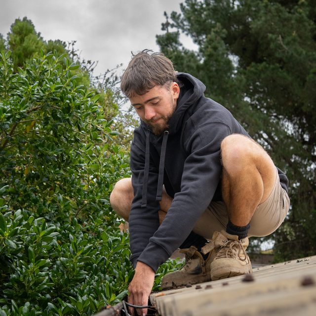 Gutter cleaner crouched on a roof edge clearing leaves, trees behind